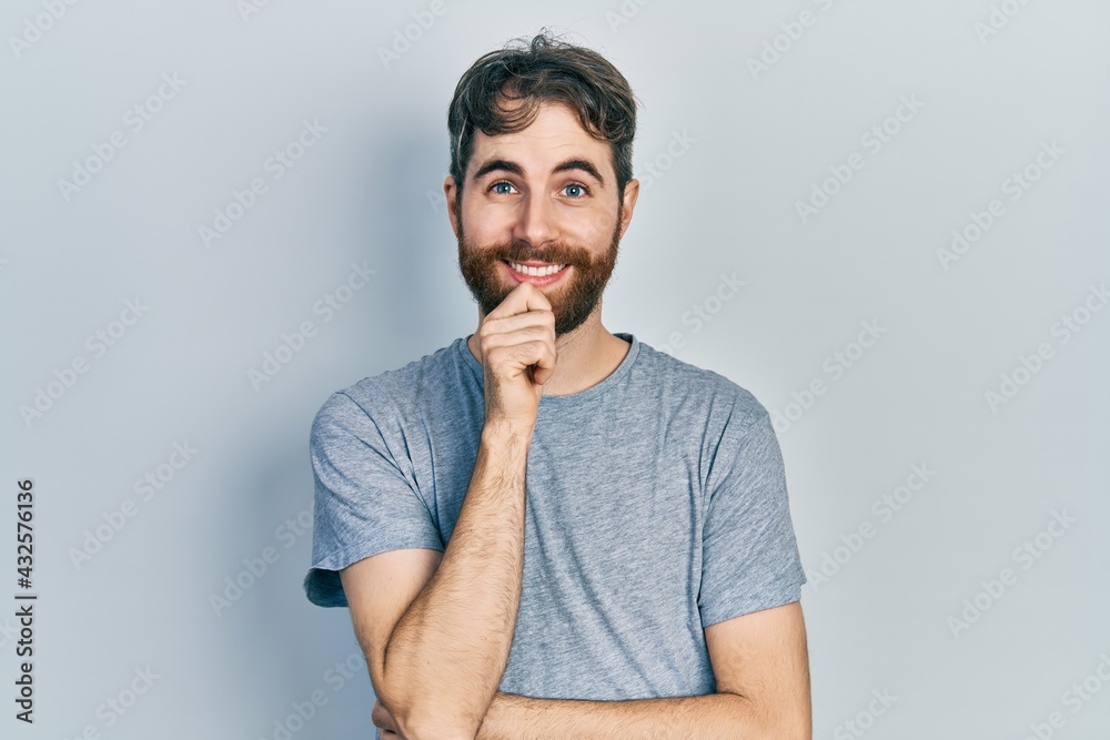 Caucasian man with beard wearing casual grey t shirt looking confident at the camera with smile with crossed arms and hand raised on chin. thinking positive. Caucasian man with beard wearing casual grey t shirt looking confident at the camera with smile with crossed arms and hand raised on chin. thinking positive.