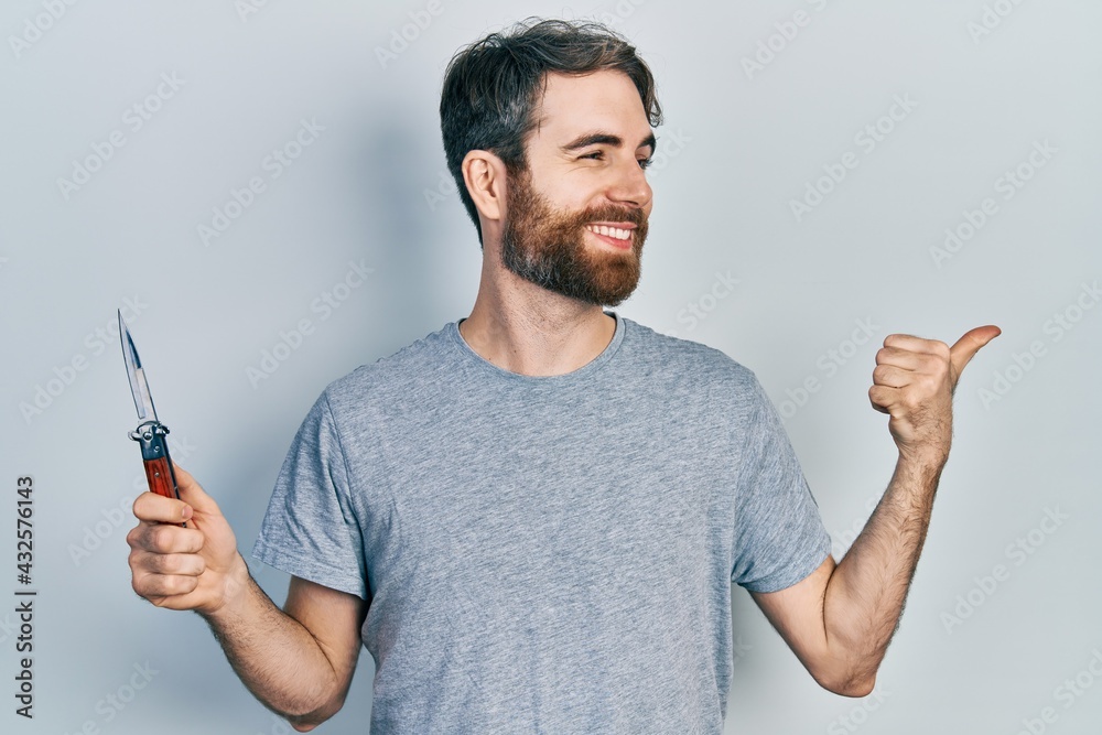Caucasian man with beard holding pocket knife pointing thumb up to the side smiling happy with open mouth Caucasian man with beard holding pocket knife pointing thumb up to the side smiling happy with open mouth