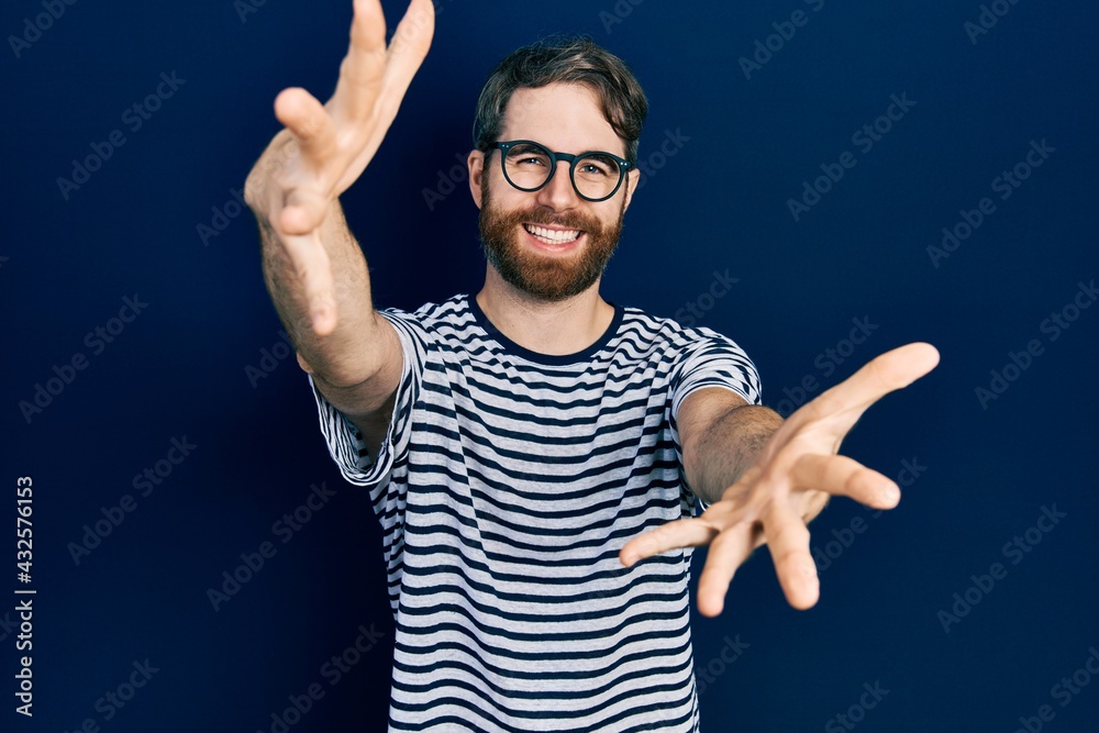 Naklejka premium Caucasian man with beard wearing striped t shirt and glasses looking at the camera smiling with open arms for hug. cheerful expression embracing happiness.