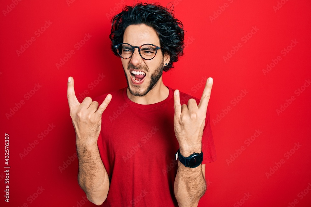 Handsome hispanic man wearing casual t shirt and glasses shouting with crazy expression doing rock symbol with hands up. music star. heavy concept. Handsome hispanic man wearing casual t shirt and glasses shouting with crazy expression doing rock symbol with hands up. music star. heavy concept.