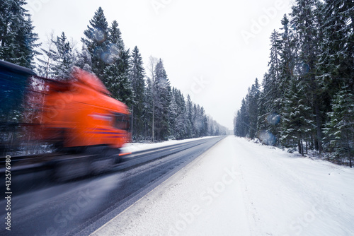 The truck is driving along the winter road during the snowfall passing through the spruce forest. View from the side of the road, image in the blue toning © Georgii Shipin