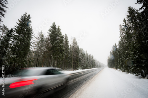 The car goes along the winter road during the snowfall passing through the spruce forest. View from the side of the road, image in the blue toning