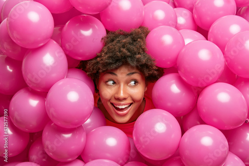 Beautiful happy curly dark skinned woman sticks head through heap of pink inflated balloons smiles joyfully looks away has festive mood enjoys party celebration Fototapet