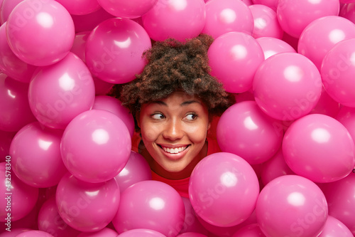 Happy cute young African American woman with curly bushy hair smiles broadly looks right has festive mood poses around inflated helium pink balloons expresses positive emotions. Holiday decor.