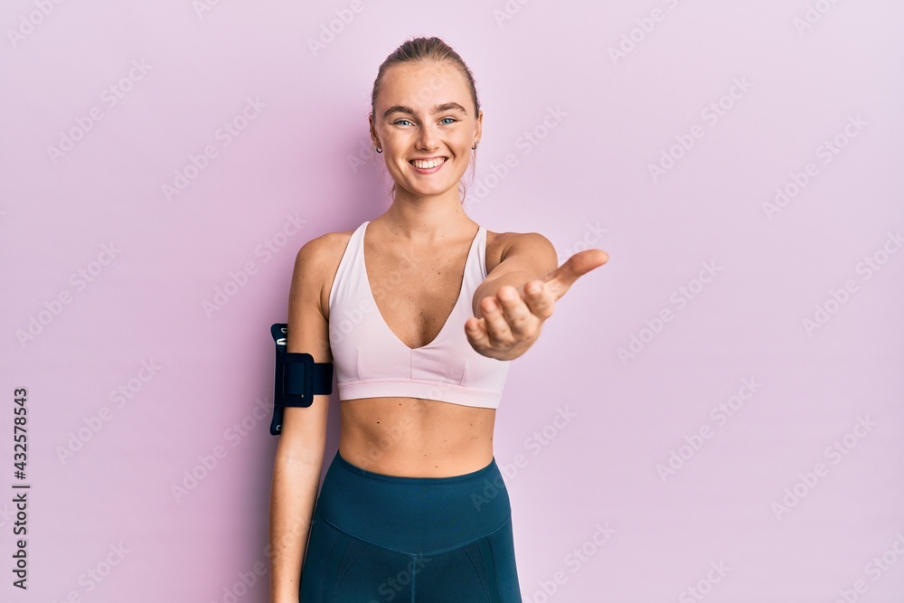 Beautiful blonde woman wearing sportswear and arm band smiling friendly offering handshake as greeting and welcoming. successful business.