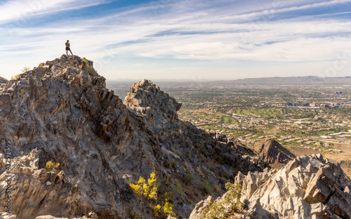фотографія A lone hiker is seen atop Piestewa Peak in Phoenix, Arizona, on top of a rocky cliff overlooking the Valley of the Sun with a blue sky with high clouds