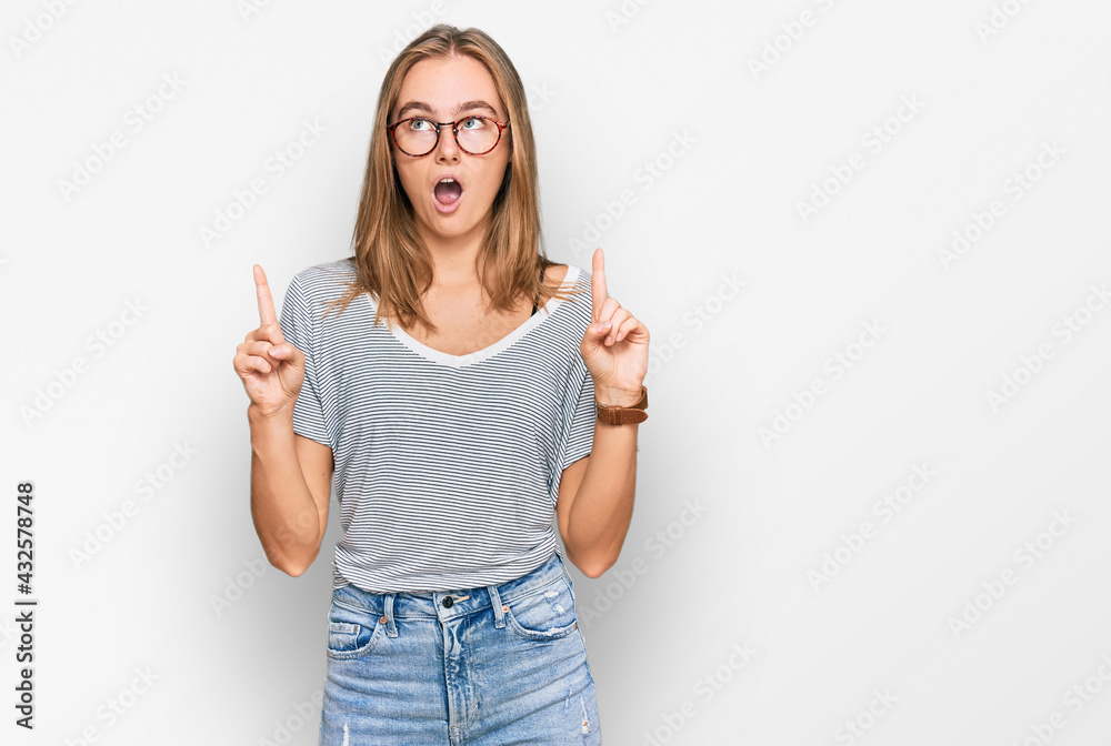 Beautiful young blonde woman wearing casual clothes and glasses amazed and surprised looking up and pointing with fingers and raised arms. Beautiful young blonde woman wearing casual clothes and glasses amazed and surprised looking up and pointing with fingers and raised arms.