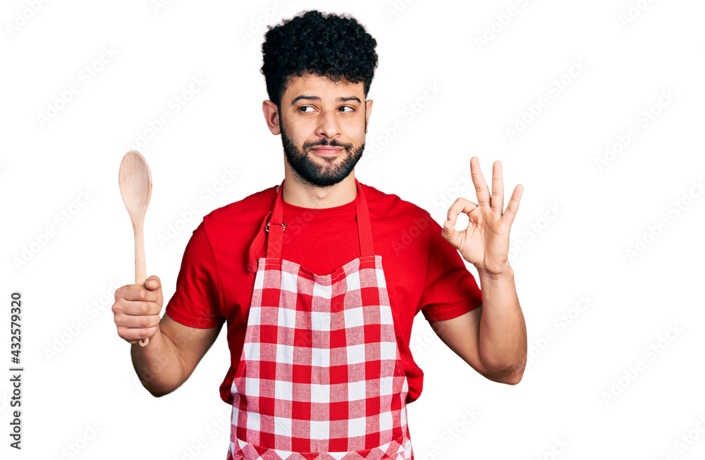 Young arab man with beard wearing baker uniform holding wooden spoon smiling looking to the side and staring away thinking. Young arab man with beard wearing baker uniform holding wooden spoon smiling looking to the side and staring away thinking.