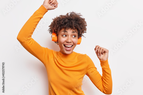 Positive dark skinned young woman with curly hair raises arms and dances carefree wears casual jumper smiles broadly catches every bit of music moves with rhythm isolated over white background