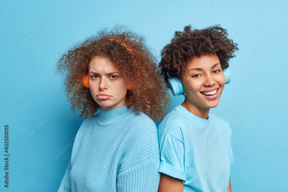 © WHstudio Leushin N - Indoor shot of displeased curly haired woman and her best friend companion stand back to each other listen music in wireless headphones isolated over blue background. Diverse women pose closely © WHstudio Leushin N - Indoor shot of displeased curly haired woman and her best friend companion stand back to each other listen music in wireless headphones isolated over blue background. Diverse women pose closely