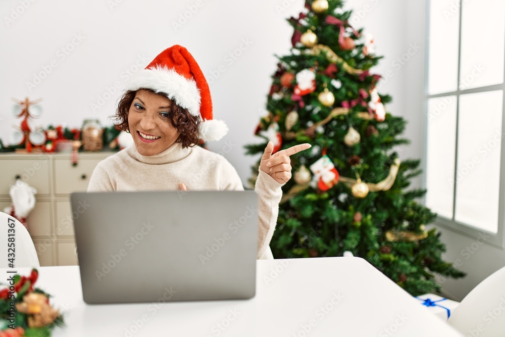 Middle age hispanic woman using laptop sitting by christmas tree smiling and looking at the camera pointing with two hands and fingers to the side. Middle age hispanic woman using laptop sitting by christmas tree smiling and looking at the camera pointing with two hands and fingers to the side.