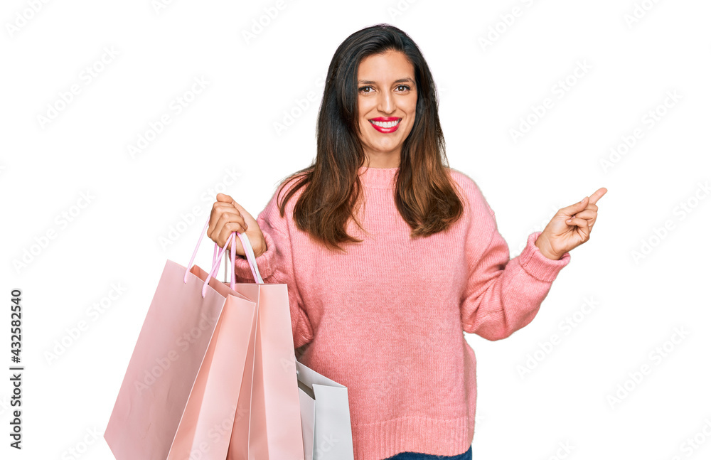 Beautiful hispanic woman holding shopping bags smiling happy pointing with hand and finger to the side Beautiful hispanic woman holding shopping bags smiling happy pointing with hand and finger to the side