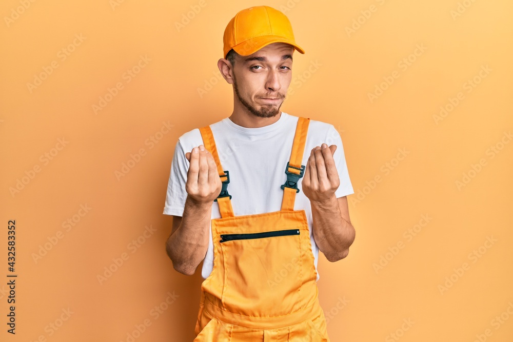Hispanic young man wearing handyman uniform doing money gesture with hands, asking for salary payment, millionaire business