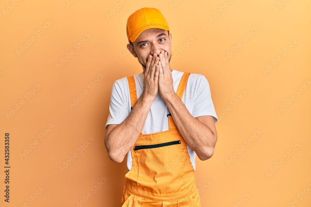 Hispanic young man wearing handyman uniform laughing and embarrassed giggle covering mouth with hands, gossip and scandal concept Hispanic young man wearing handyman uniform laughing and embarrassed giggle covering mouth with hands, gossip and scandal concept