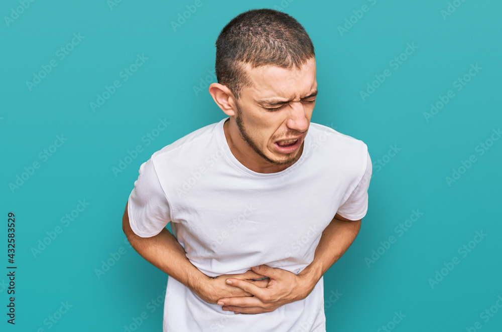 Hispanic young man wearing casual white t shirt with hand on stomach because nausea, painful disease feeling unwell. ache concept. Hispanic young man wearing casual white t shirt with hand on stomach because nausea, painful disease feeling unwell. ache concept.