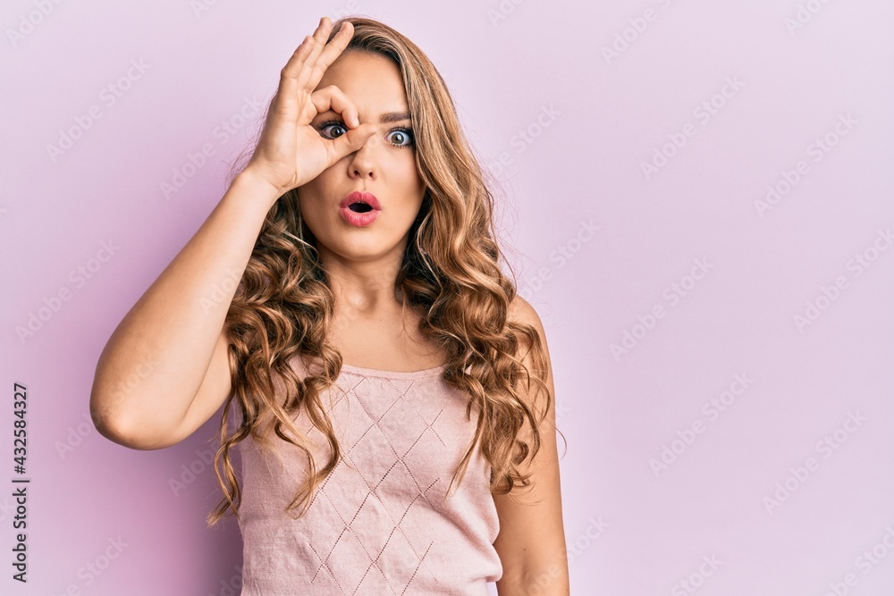 Young blonde girl wearing casual clothes doing ok gesture shocked with surprised face, eye looking through fingers. unbelieving expression.