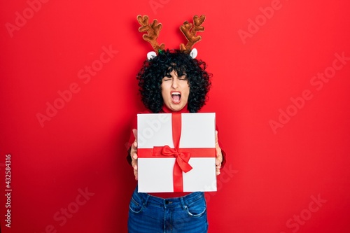 Young middle east woman wearing deer christmas hat holding gift angry and mad screaming frustrated and furious, shouting with anger looking up.