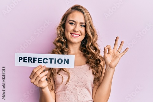 Young blonde girl holding paper with perfection message doing ok sign with fingers, smiling friendly gesturing excellent symbol