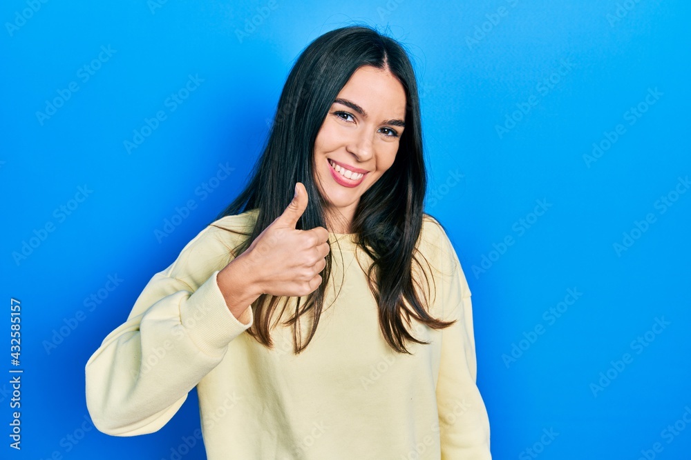 Young brunette woman wearing casual sweatshirt doing happy thumbs up gesture with hand. approving expression looking at the camera showing success. Young brunette woman wearing casual sweatshirt doing happy thumbs up gesture with hand. approving expression looking at the camera showing success.
