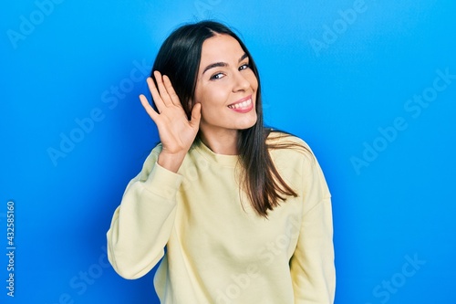 Young brunette woman wearing casual sweatshirt smiling with hand over ear listening an hearing to rumor or gossip. deafness concept.