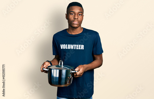 Young african american man wearing volunteer holding cooking pot relaxed with serious expression on face. simple and natural looking at the camera.