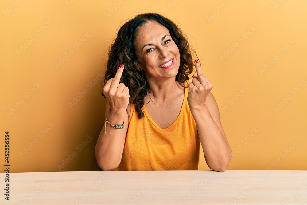 Beautiful middle age woman wearing casual clothes sitting on the table showing middle finger doing fuck you bad expression, provocation and rude attitude. screaming excited