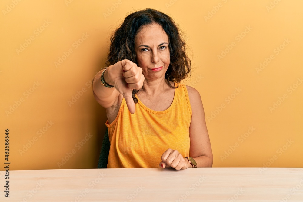 Beautiful middle age woman wearing casual clothes sitting on the table looking unhappy and angry showing rejection and negative with thumbs down gesture. bad expression. Beautiful middle age woman wearing casual clothes sitting on the table looking unhappy and angry showing rejection and negative with thumbs down gesture. bad expression.