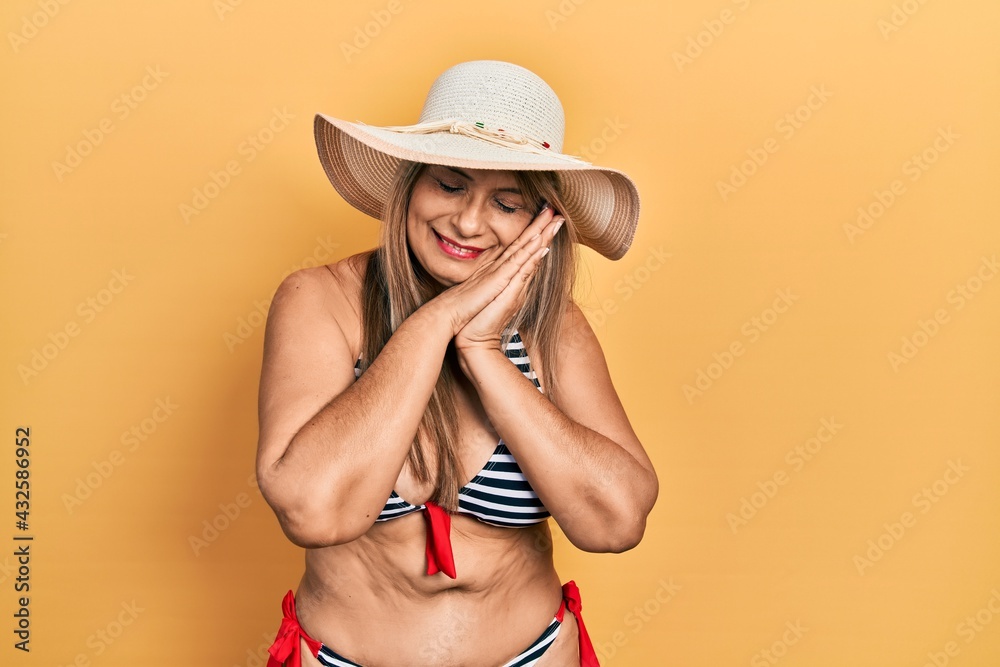 Middle age hispanic woman wearing bikini and summer hat sleeping tired dreaming and posing with hands together while smiling with closed eyes. Middle age hispanic woman wearing bikini and summer hat sleeping tired dreaming and posing with hands together while smiling with closed eyes.