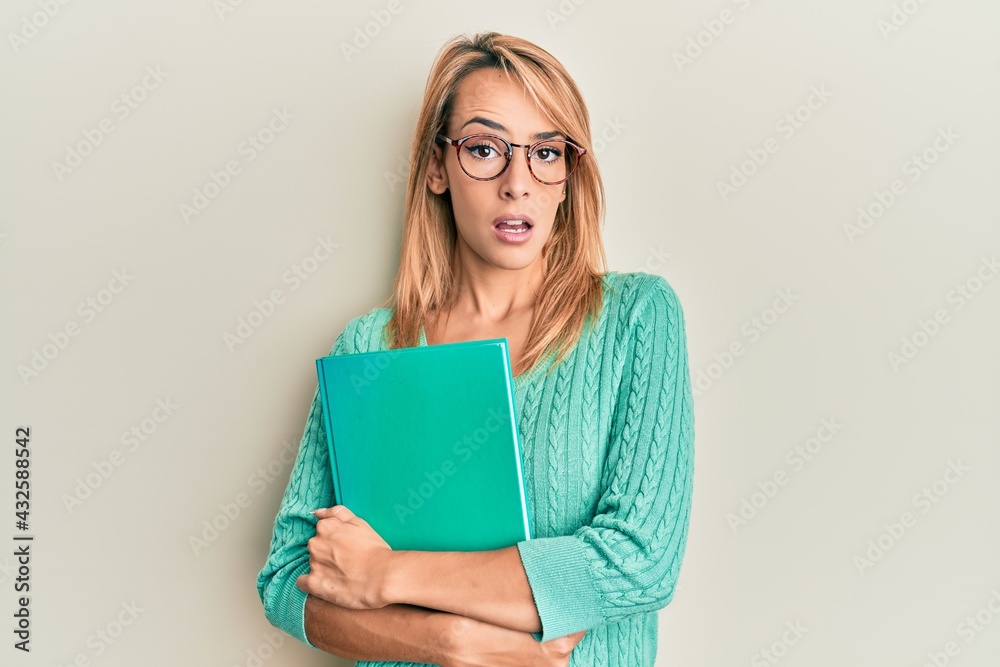 Beautiful blonde woman holding book wearing glasses in shock face, looking skeptical and sarcastic, surprised with open mouth Beautiful blonde woman holding book wearing glasses in shock face, looking skeptical and sarcastic, surprised with open mouth
