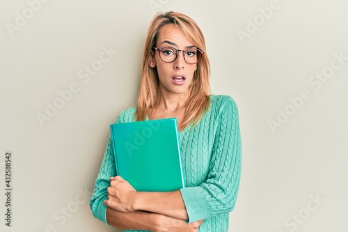 Beautiful blonde woman holding book wearing glasses in shock face, looking skeptical and sarcastic, surprised with open mouth