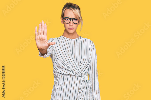 Beautiful blonde woman wearing business shirt and glasses doing stop sing with palm of the hand. warning expression with negative and serious gesture on the face.