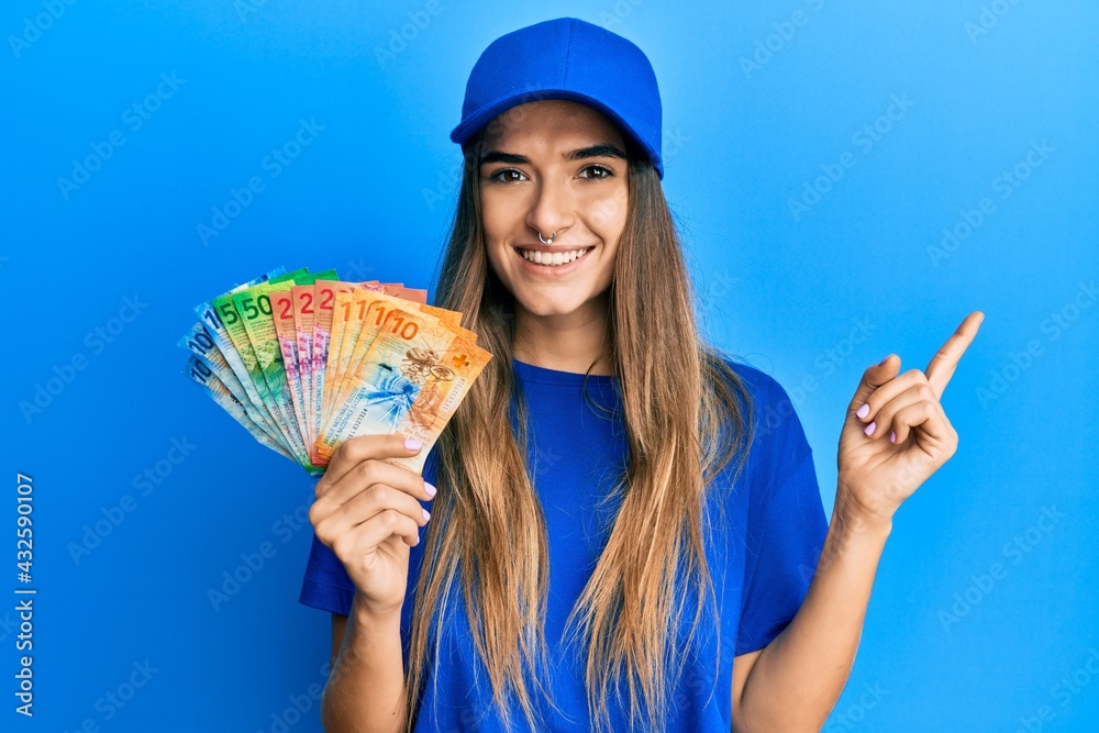 Young hispanic woman wearing delivery uniform and cap holding swiss franc smiling happy pointing with hand and finger to the side Young hispanic woman wearing delivery uniform and cap holding swiss franc smiling happy pointing with hand and finger to the side