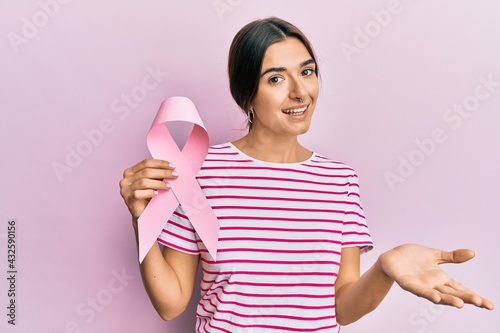 Young hispanic woman holding pink cancer ribbon celebrating achievement with happy smile and winner expression with raised hand