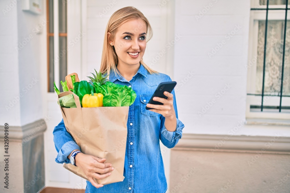 Young blonde girl smiling happy using smartphone and holding grocries paper bag at the city. Young blonde girl smiling happy using smartphone and holding grocries paper bag at the city.