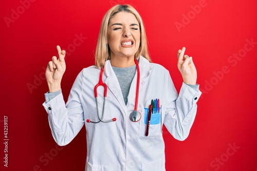 Young caucasian woman wearing doctor uniform and stethoscope gesturing finger crossed smiling with hope and eyes closed. luck and superstitious concept.