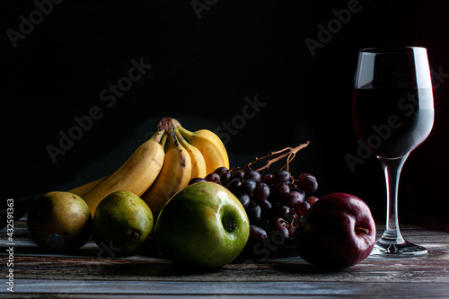 Baroque, renaissance still life fruits on a wooden rustic table with a glass of wine on the side with bananas grapes pears red apple and green apple low key light chiaroscuro dark food photo