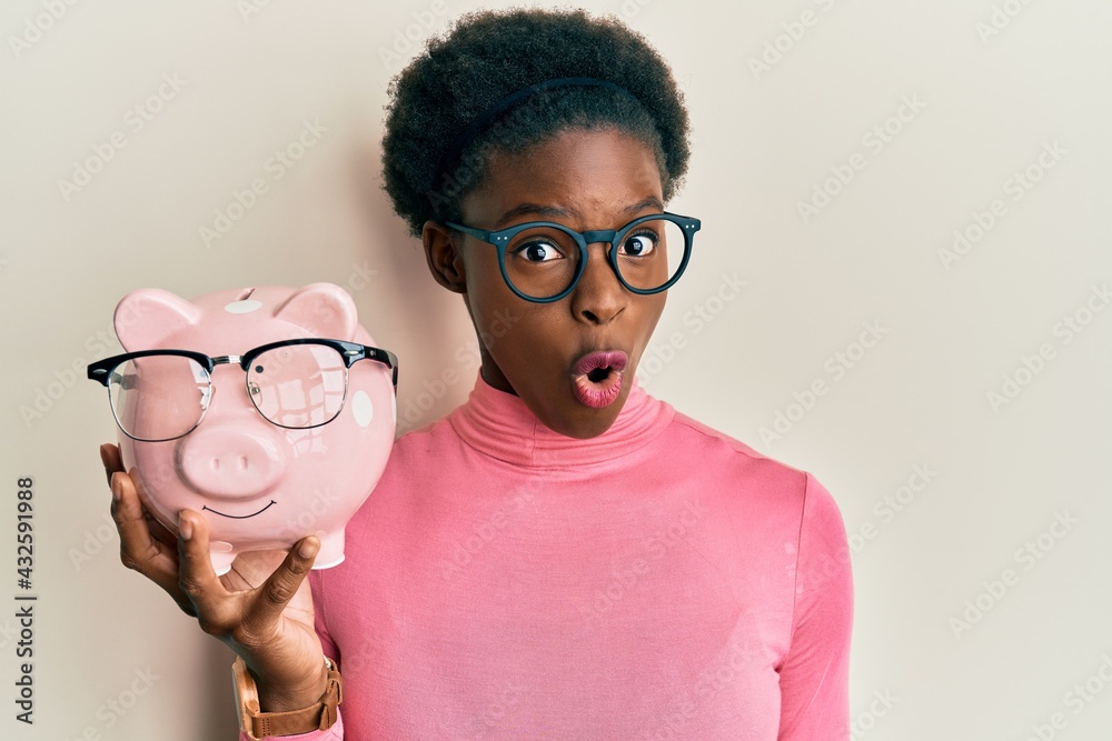 Young african american girl holding piggy bank with glasses scared and amazed with open mouth for surprise, disbelief face Young african american girl holding piggy bank with glasses scared and amazed with open mouth for surprise, disbelief face