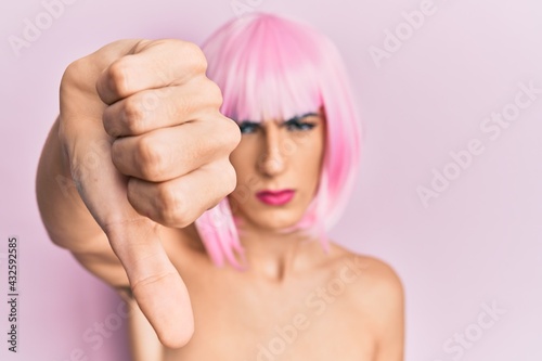 Young man wearing woman make up wearing pink wig looking unhappy and angry showing rejection and negative with thumbs down gesture. bad expression.