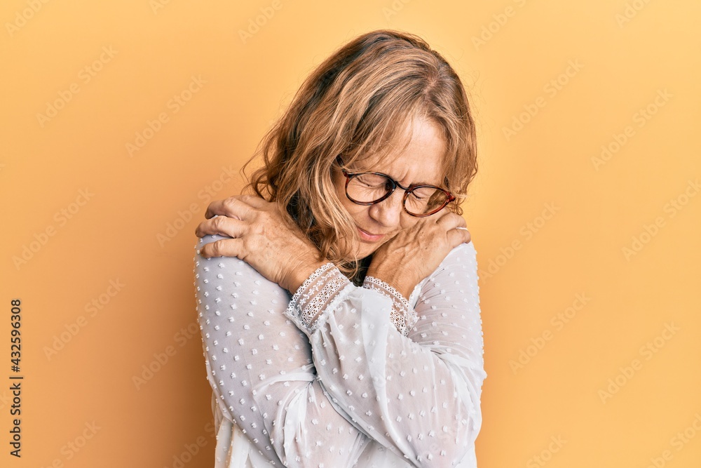 Middle age blonde woman wearing casual clothes over yellow background hugging oneself happy and positive, smiling confident. self love and self care