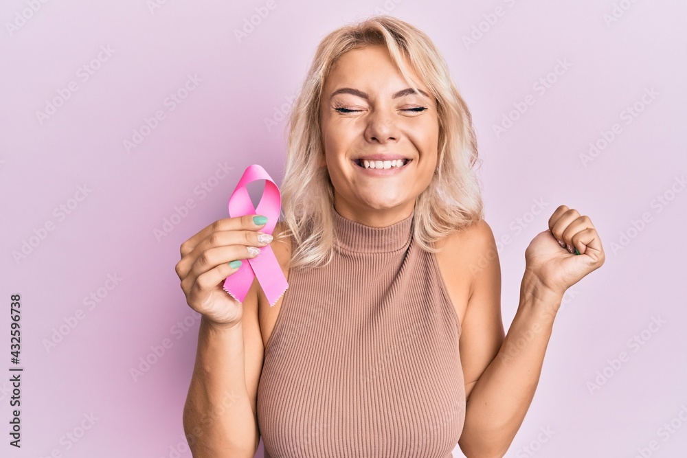 Young blonde girl holding pink cancer ribbon screaming proud, celebrating victory and success very excited with raised arm