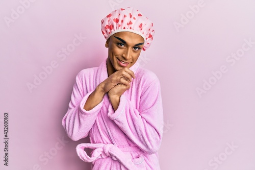 Hispanic man wearing make up wearing shower towel cap and bathrobe laughing nervous and excited with hands on chin looking to the side