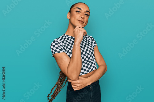 Hispanic transgender man wearing make up and long hair wearing modern clothes looking confident at the camera with smile with crossed arms and hand raised on chin. thinking positive.