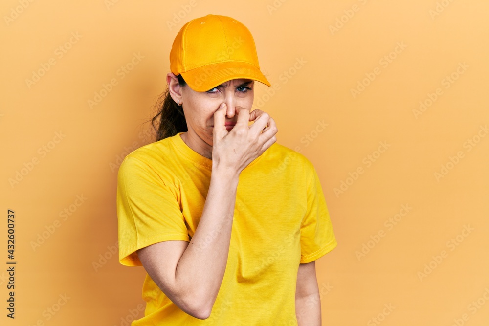 Young hispanic woman wearing delivery uniform and cap smelling something stinky and disgusting, intolerable smell, holding breath with fingers on nose. bad smell