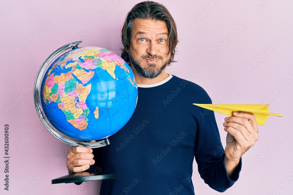 Fototapeta premium Middle age handsome man holding paper plane and world ball puffing cheeks with funny face. mouth inflated with air, catching air.
