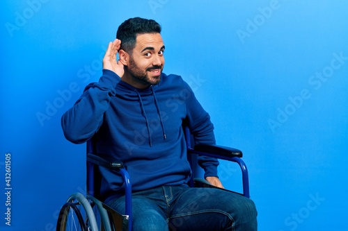 Handsome hispanic man with beard sitting on wheelchair smiling with hand over ear listening an hearing to rumor or gossip. deafness concept.