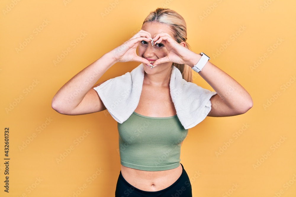 Young blonde girl wearing sportswear and towel doing heart shape with hand and fingers smiling looking through sign Young blonde girl wearing sportswear and towel doing heart shape with hand and fingers smiling looking through sign