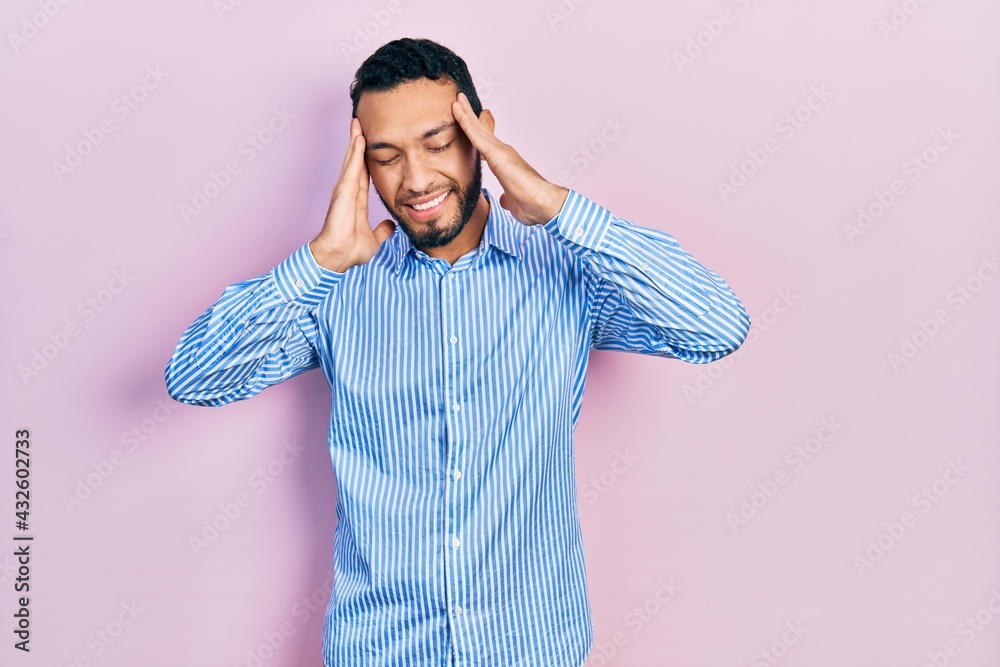 Hispanic man with beard wearing casual blue shirt with hand on head for pain in head because stress. suffering migraine. Hispanic man with beard wearing casual blue shirt with hand on head for pain in head because stress. suffering migraine.