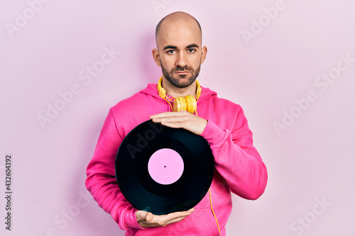 Young bald man holding vinyl disc relaxed with serious expression on face. simple and natural looking at the camera.