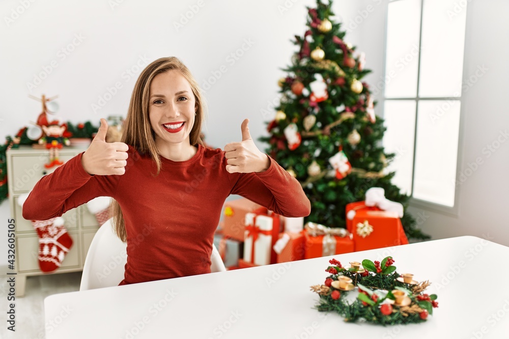 Caucasian young blonde woman sitting on the table by christmas tree success sign doing positive gesture with hand, thumbs up smiling and happy. cheerful expression and winner gesture. Caucasian young blonde woman sitting on the table by christmas tree success sign doing positive gesture with hand, thumbs up smiling and happy. cheerful expression and winner gesture.