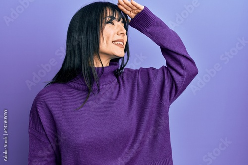 Young brunette woman with bangs wearing turtleneck sweater very happy and smiling looking far away with hand over head. searching concept. © Krakenimages.com